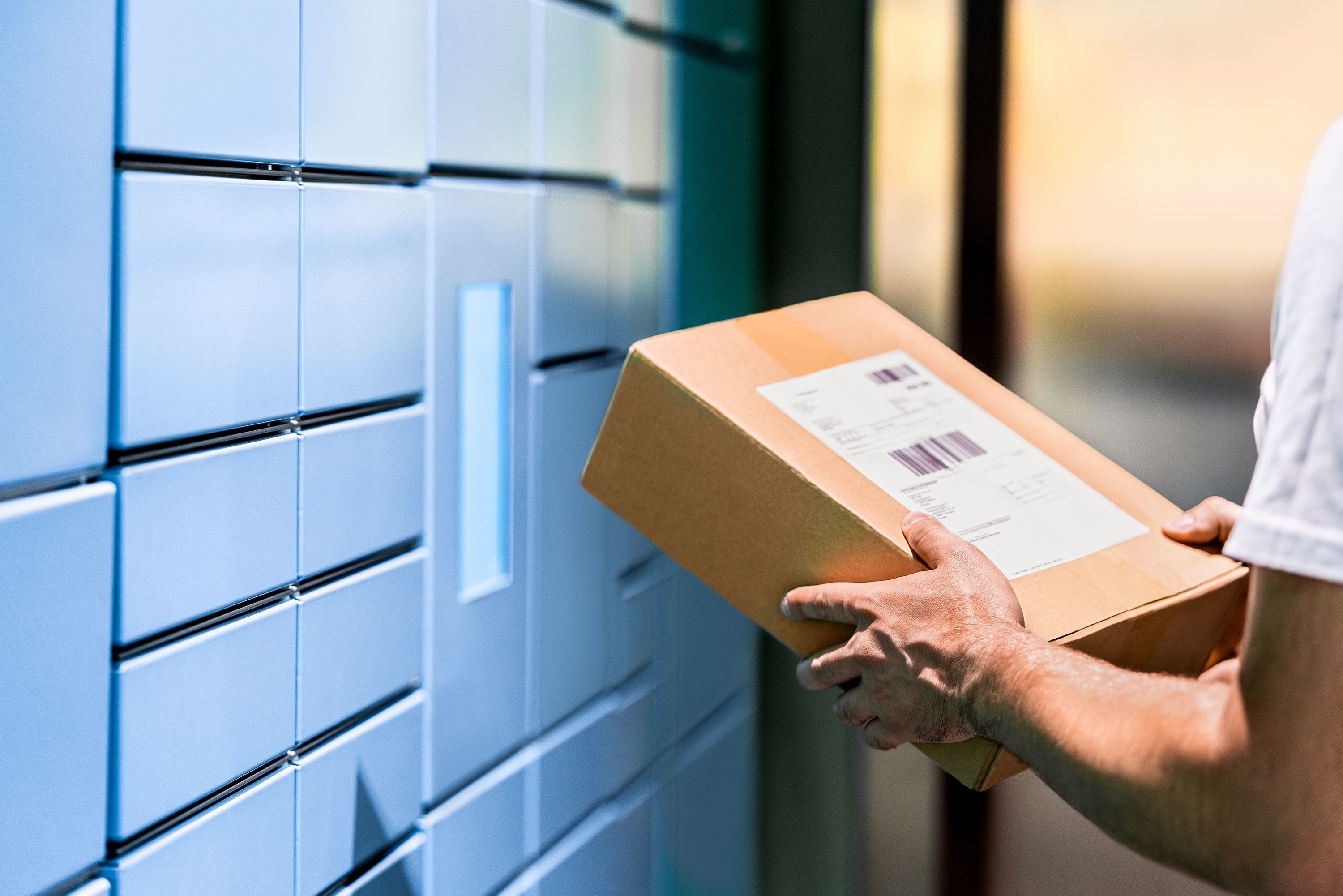 person holding a small brown box in front of blue package mailboxes